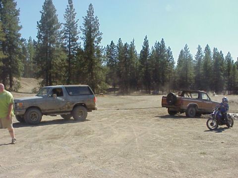 1989 Ford Bronco - Two Ford Broncos are in a dusty outdoor area, one towing the other, with a dirt bike in the foreground.