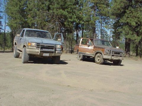1989 Ford Bronco - Two Ford Broncos, one light blue and one brown, are parked on a dirt road with trees in the background.