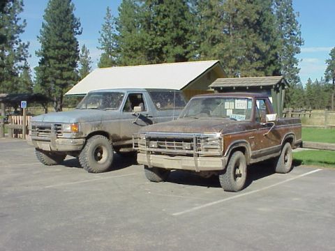 1989 Ford Bronco - A 1989 Ford Bronco and a brown pickup truck are parked in a lot next to a building.