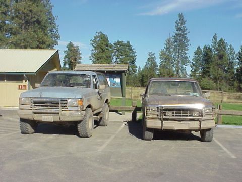 1989 Ford Bronco - Two Ford Broncos are parked side-by-side in an outdoor lot with trees and a building in the background.