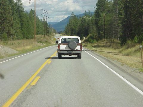 1989 Ford Bronco - A brown and white Ford Bronco is seen from behind on a two-lane highway, with mountains and trees in the background.