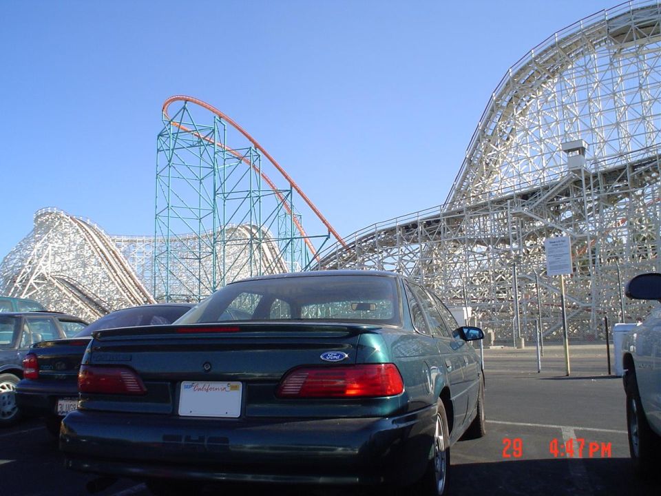 Green Ford SHO sedan rear at an amusement park with roller coasters behind