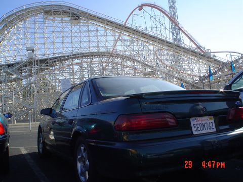 1993 Ford Taurus - A dark sedan is parked in front of a roller coaster at an amusement park. The car's model year is visible.
