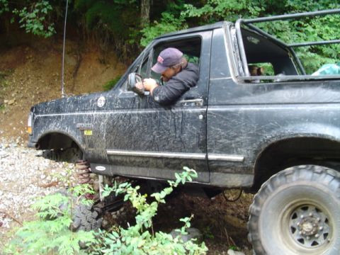 1993 Ford Bronco - A driver maneuvers a muddy black 1993 Ford Bronco through an off-road trail.
