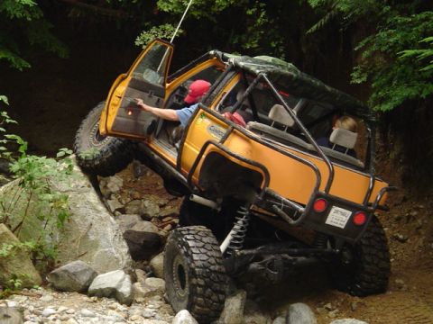 1993 Ford Bronco - A modified yellow off-road vehicle navigates a challenging rocky terrain on a wooded trail.