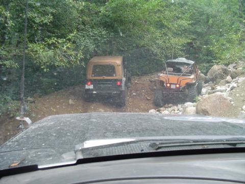 1993 Ford Bronco - Two off-road vehicles navigate a rocky and uneven dirt trail surrounded by dense foliage.