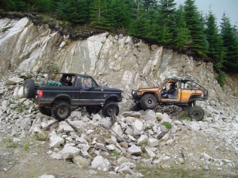 1993 Ford Bronco - Two modified off-road vehicles navigate a challenging rocky incline with steep cliffs and trees in the background.