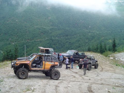 1993 Ford Bronco - A group of modified off-road vehicles are gathered on a mountain trail, with a dense forest and misty clouds in the background.