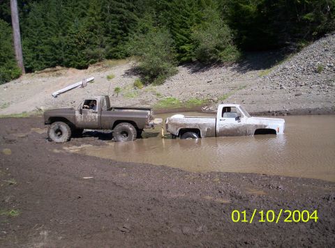 1983 Ford Bronco - Two pickup trucks are stuck in a muddy area with trees in the background.