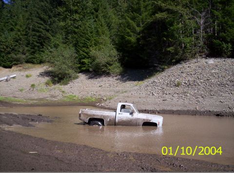 1983 Ford Bronco - A white pickup truck is partially submerged in muddy water, with a wooded hillside behind it.