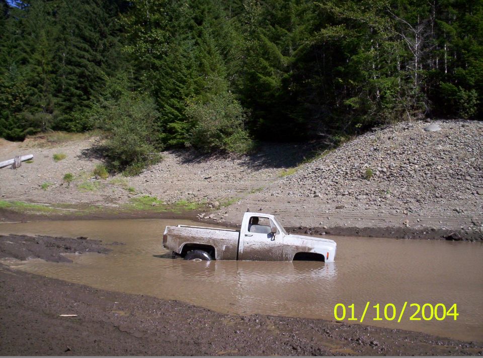 White pickup truck stuck in muddy water with trees and a rocky hill in the background