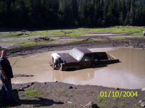 1983 Ford Bronco - A pickup truck buried to its door tops in a brown mud pit, with a man in a white shirt wading in to hook a recovery strap to the front of t…