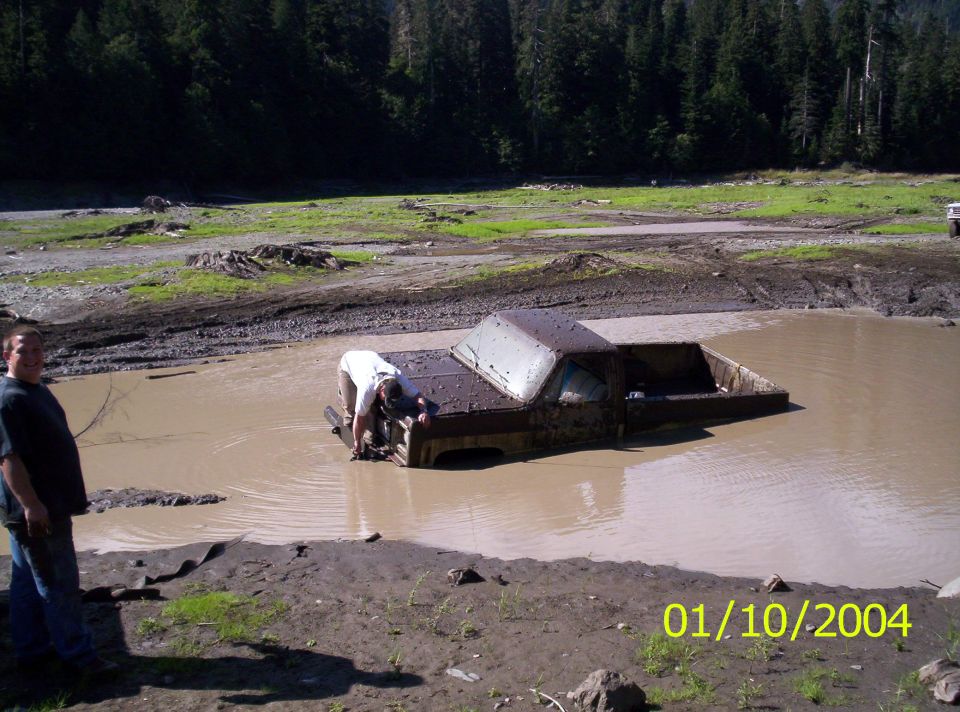 Pickup truck sunk to the doors in a muddy pit while a man wades in to hook a strap