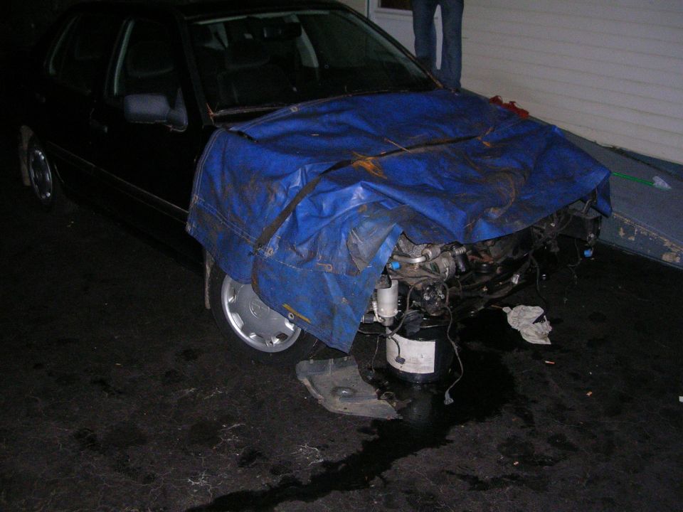 Black sedan with front end damage covered by a blue tarp, engine exposed