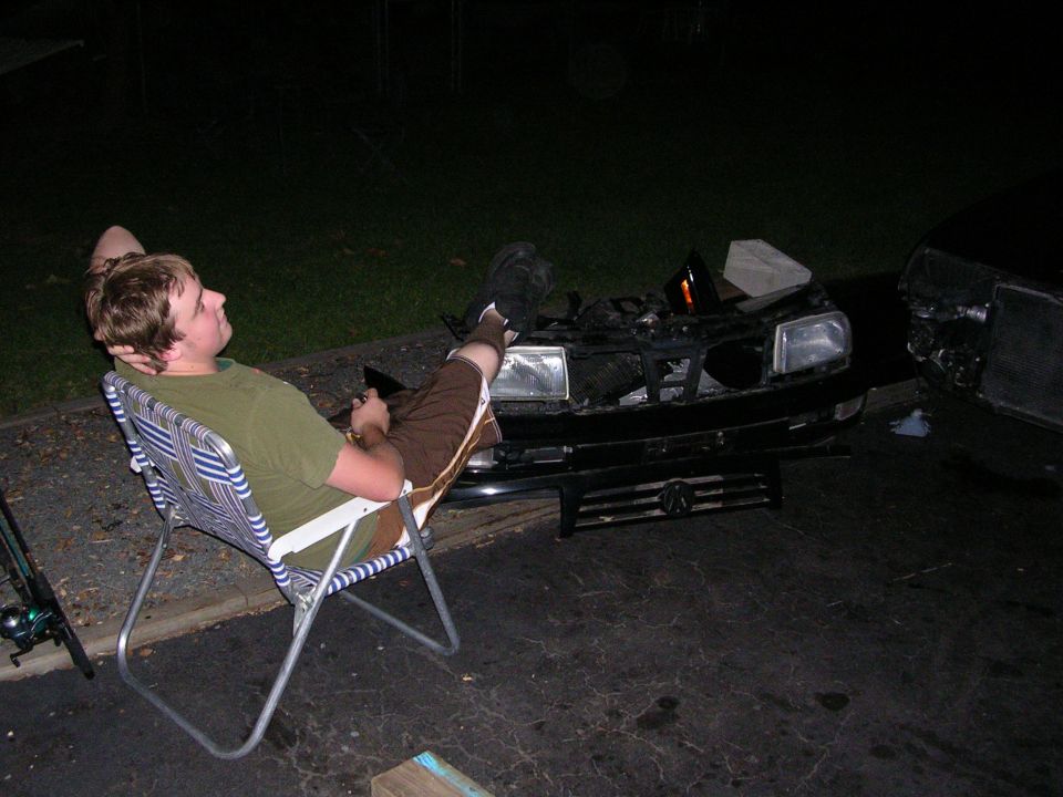 Person relaxing in a lawn chair beside the front end of a car with its hood open
