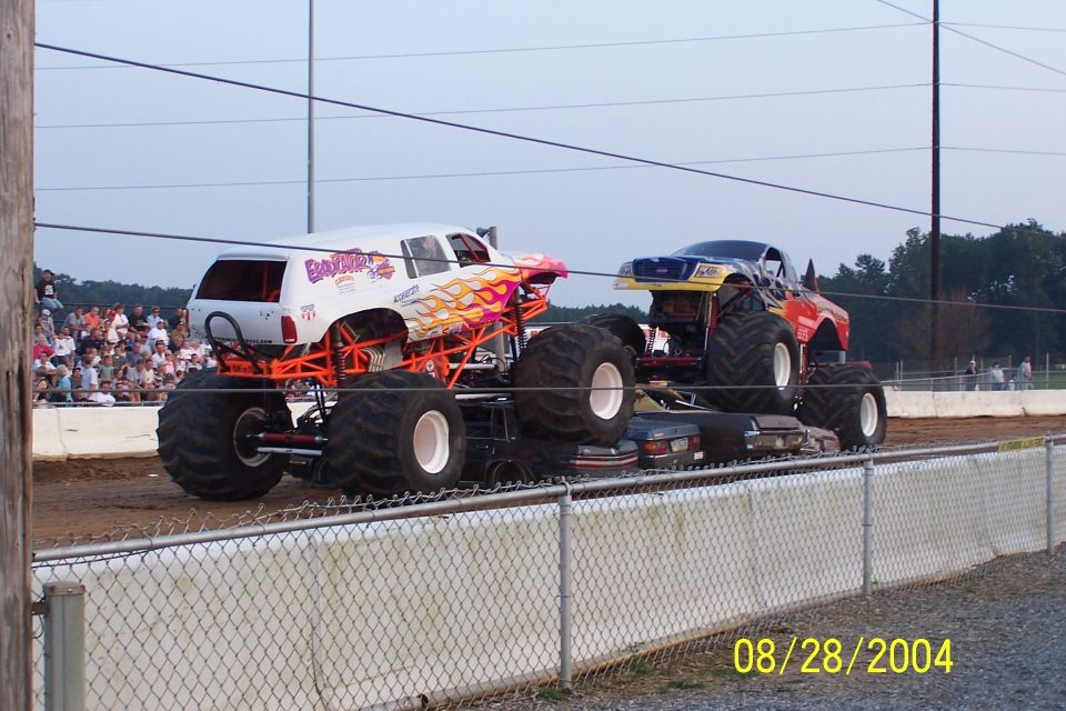Two monster trucks, one white with flames and "Banzai" text, the other red and blue, crushing cars