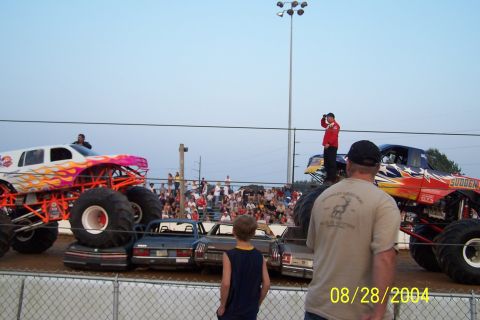 1979 Ford Bronco II - Two monster trucks, one white with flames and one red with stars, are positioned over crushed cars with spectators in the background.