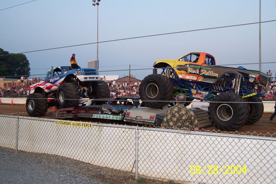 Two monster trucks, one with American flag paint and 'Bigfoot' text, the other yellow with 'Black Stallion' text, crushing cars in an arena