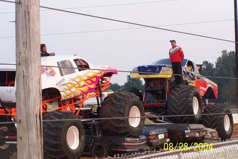1979 Ford Bronco II - Monster trucks are positioned over a pile of wrecked cars, with spectators visible in the vehicles.