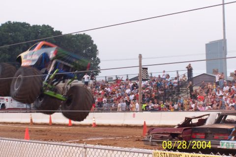 1979 Ford Bronco II - Monster truck performs a jump over a wrecked car during an event, with a crowd watching from the stands.