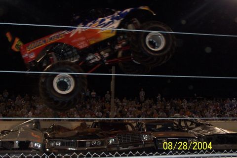 1979 Ford Bronco II - A monster truck soars over a pile of crushed cars during an event, with spectators watching.