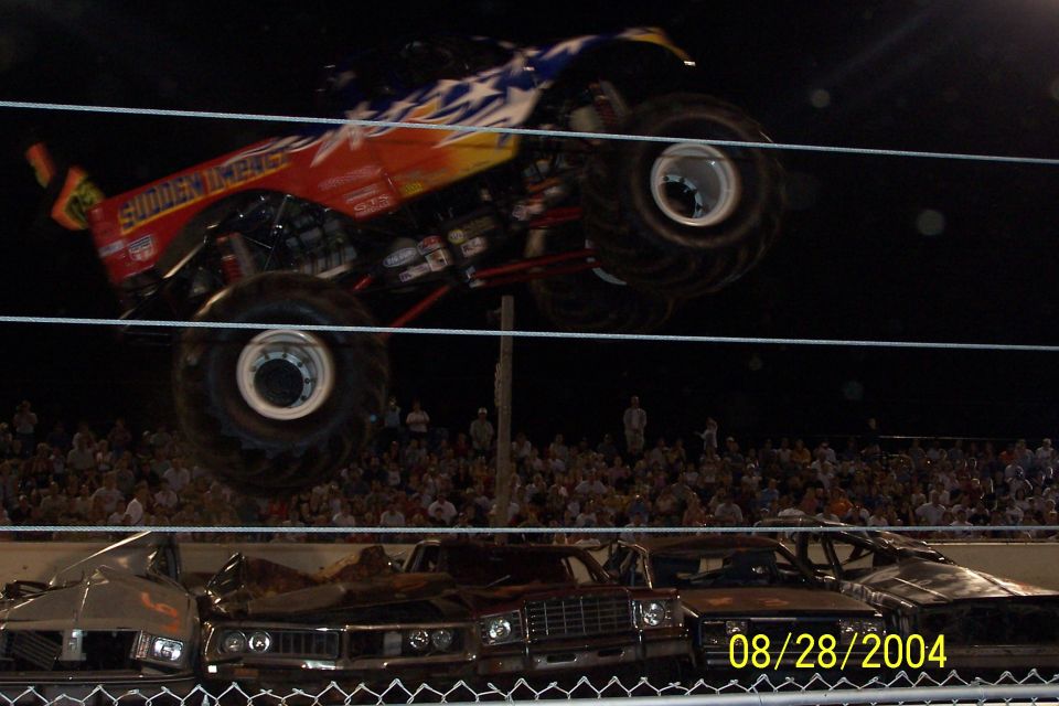 monster truck mid-air over wrecked cars, crowd in background