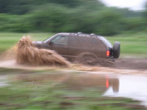 1995 Ford Bronco - A muddy SUV navigates through a water obstacle, kicking up a large spray.