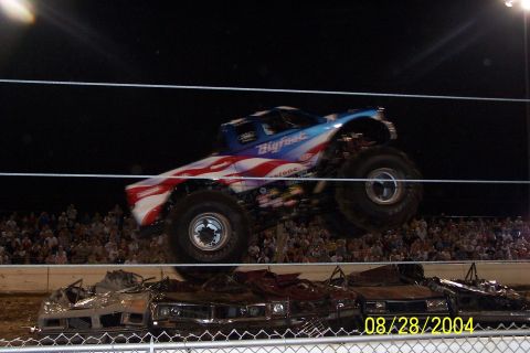 1979 Ford Bronco II - Bigfoot monster truck performs a jump over a row of wrecked cars at an event. Crowd watches from the stands.