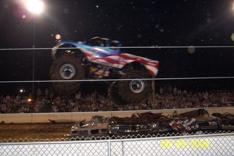 1979 Ford Bronco II - A monster truck soars through the air over a pile of wrecked cars during a nighttime event.