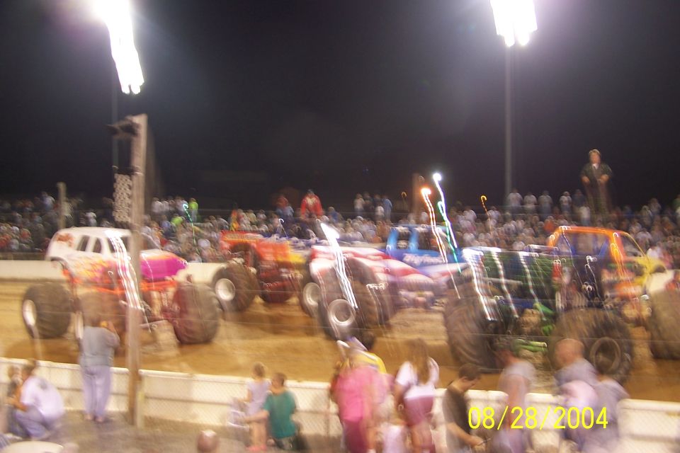 Multiple monster trucks lined up on a dirt track with crowds watching under stadium lights