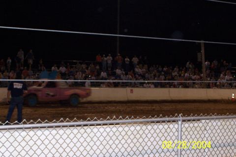 1979 Ford Bronco II - A pink pickup truck races on a dirt track at night, with a crowd watching from the stands.