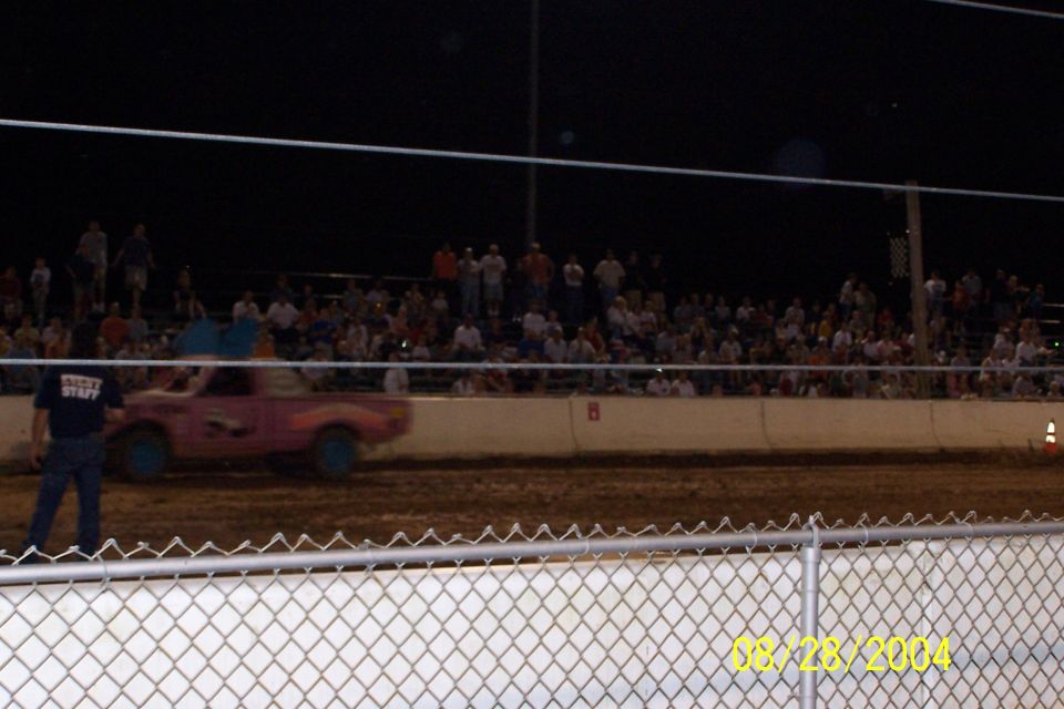 Pink pickup truck with blue wheels on a dirt track, blurred by motion, with spectators in the background