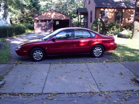 1998 Ford Taurus - Driver's-side profile of a dark red Ford Taurus sedan sitting at the end of a residential driveway, with a detached garage and tan house be…
