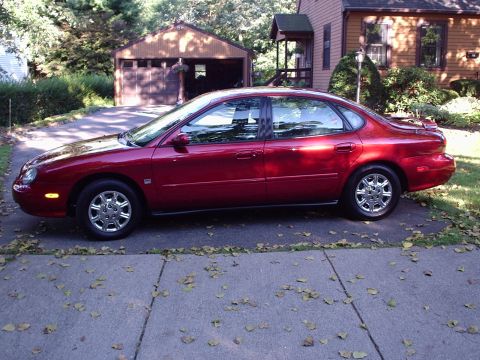 1998 Ford Taurus - Driver-side profile of a dark red four-door sedan with the rounded oval-era styling, chrome multi-spoke alloy wheels and tinted glass, park…