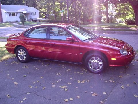 1998 Ford Taurus - A dark red Ford Taurus sedan photographed from the passenger side on a leaf-strewn suburban driveway, showing the rounded oval-era body, bo…