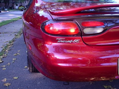 1998 Ford Taurus - Close rear-corner shot of a red Ford Taurus sedan parked at a leaf-strewn curb, framing the left tail lamp cluster with clear reverse lens…