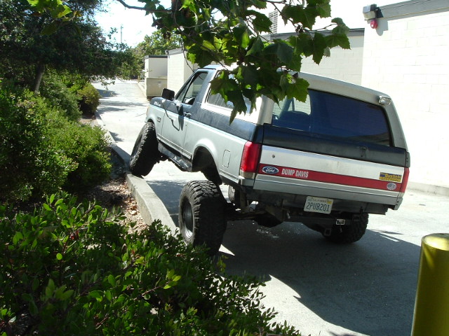 Silver lifted Ford Bronco climbing a curb, rear 3/4 view, CA plate 2PUB201.