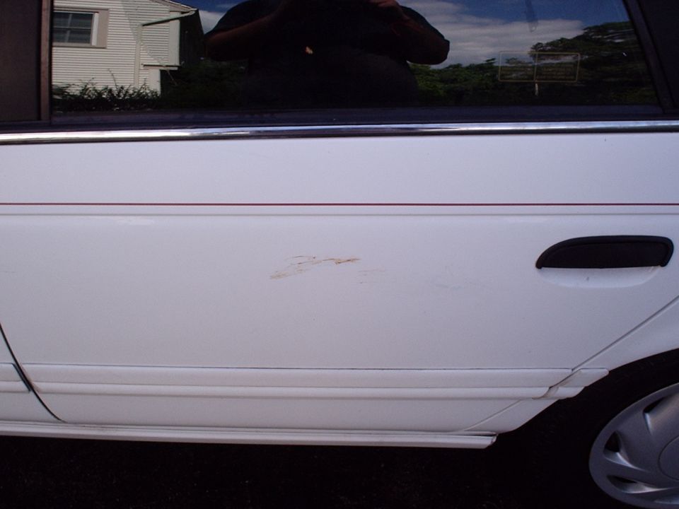White car door showing a rust spot and a black door handle. A thin red pinstripe runs horizontally above the rust