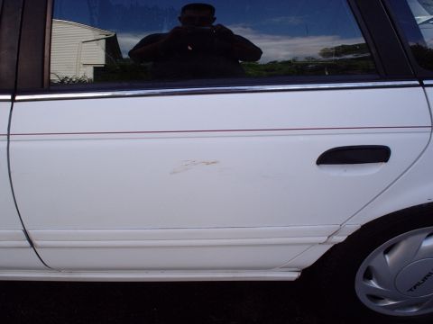 1998 Ford Taurus - The side of a white sedan is shown, with a scratch on the door and a reflection of the photographer in the window.