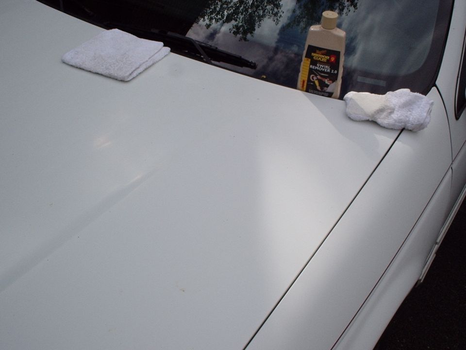 White car hood with a folded towel, a bottle of swirl remover, and a microfiber cloth