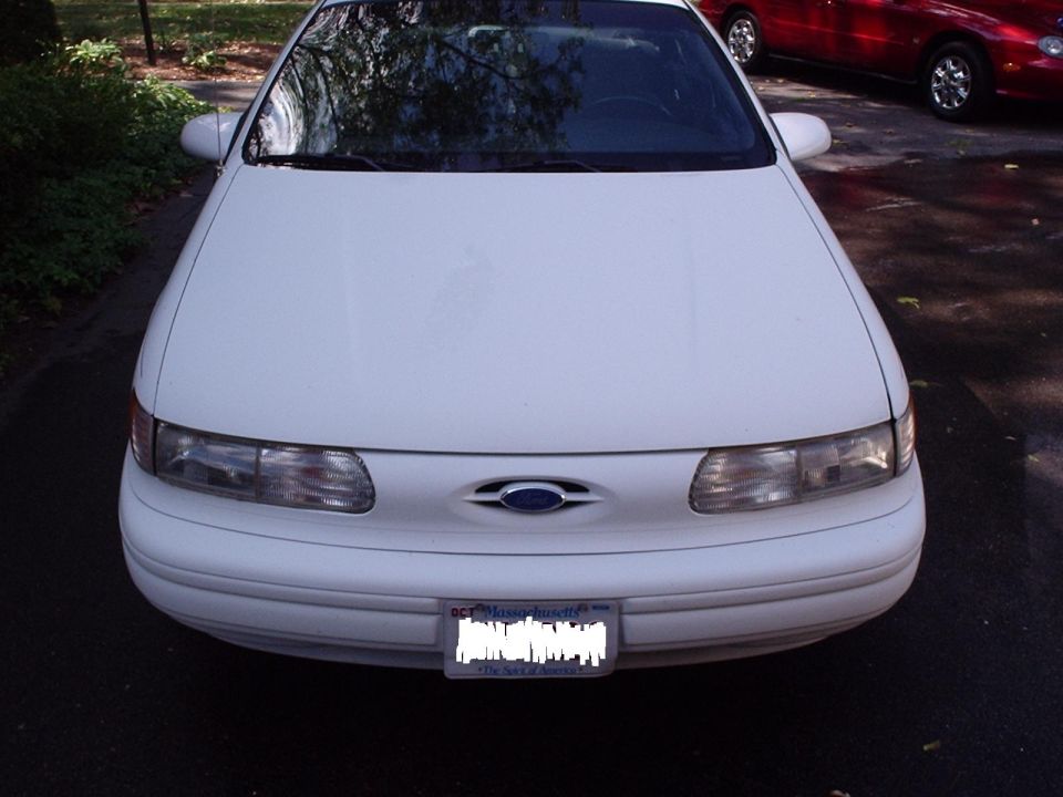 Front view of a white 1998 Ford Taurus with a Massachusetts license plate. Red car visible in background