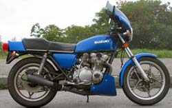 1985 Ford Crown Victoria LTD - A blue Suzuki motorcycle is shown from the side, parked outdoors with trees in the background.