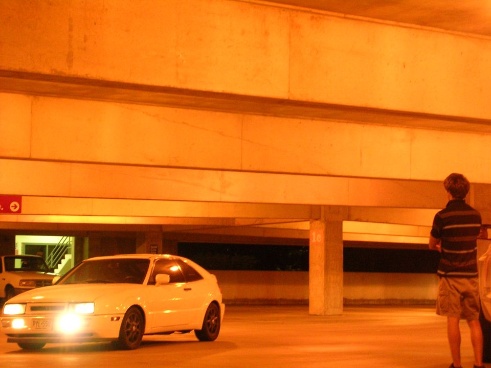 White coupe with headlights on in an orange-lit parking garage, front 3/4 view
