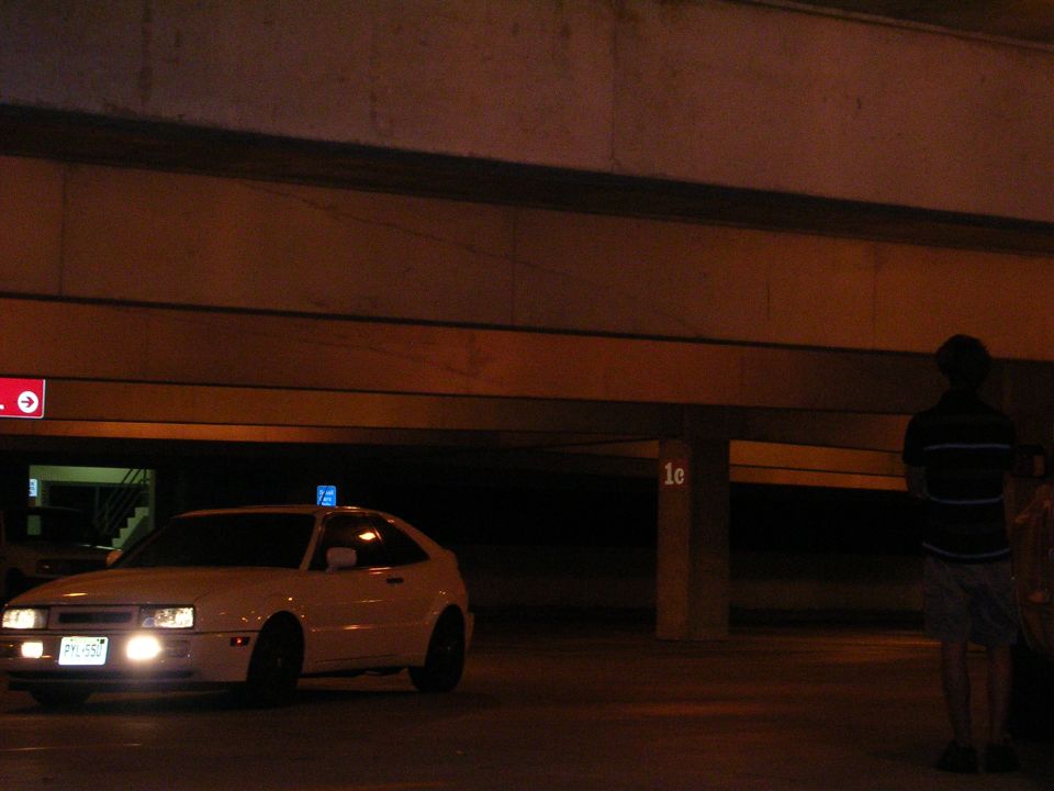 White coupe with headlights on parked in a dark concrete parking garage at night