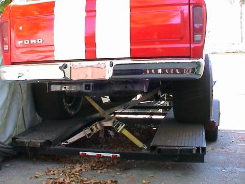 1979 Ford Bronco - The rear of a red Ford pickup truck with white racing stripes is secured to a trailer for transport.
