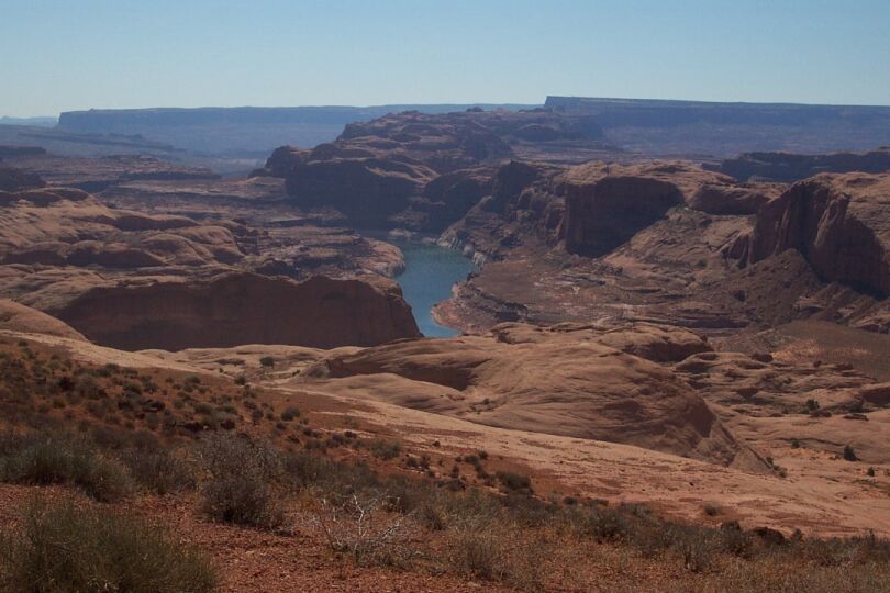 Canyon landscape with a river and red rock formations under a clear blue sky. "SUPERMOTORS" logo in the lower right corner