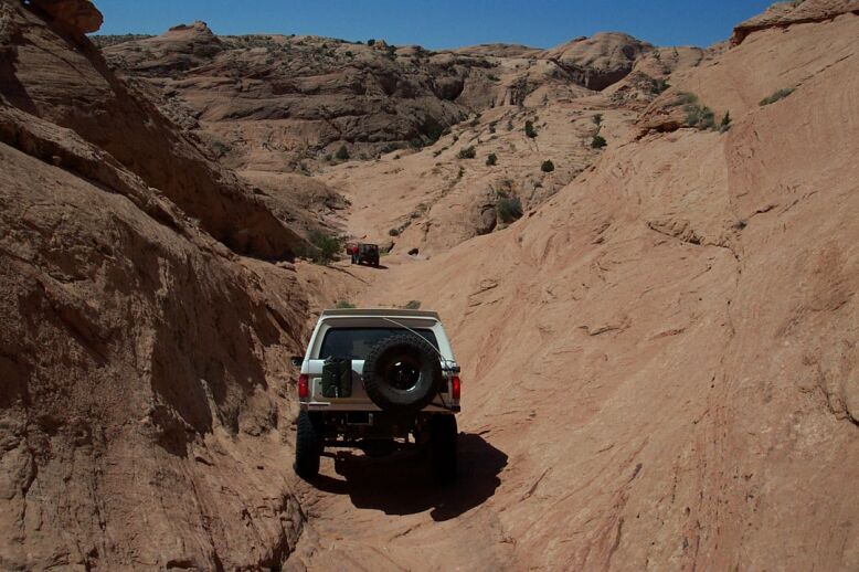 White 1990 Ford Bronco rear view, spare tire mounted, driving on rocky desert trail