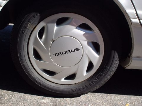 1998 Ford Taurus - Close-up of a silver swirl-spoke wheel cover stamped TAURUS on the center cap, mounted on a General AmeriGT P205/65R15 tire. The silver rea…