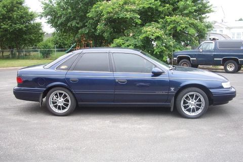 1993 Ford Taurus - A blue 1993 Ford Taurus sedan is shown from the side, aftermarket wheels and a dark blue SUV in the background.