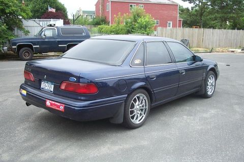 1993 Ford Taurus - A dark blue sedan is parked in a lot next to a dark blue SUV. The sedan has chrome wheels.